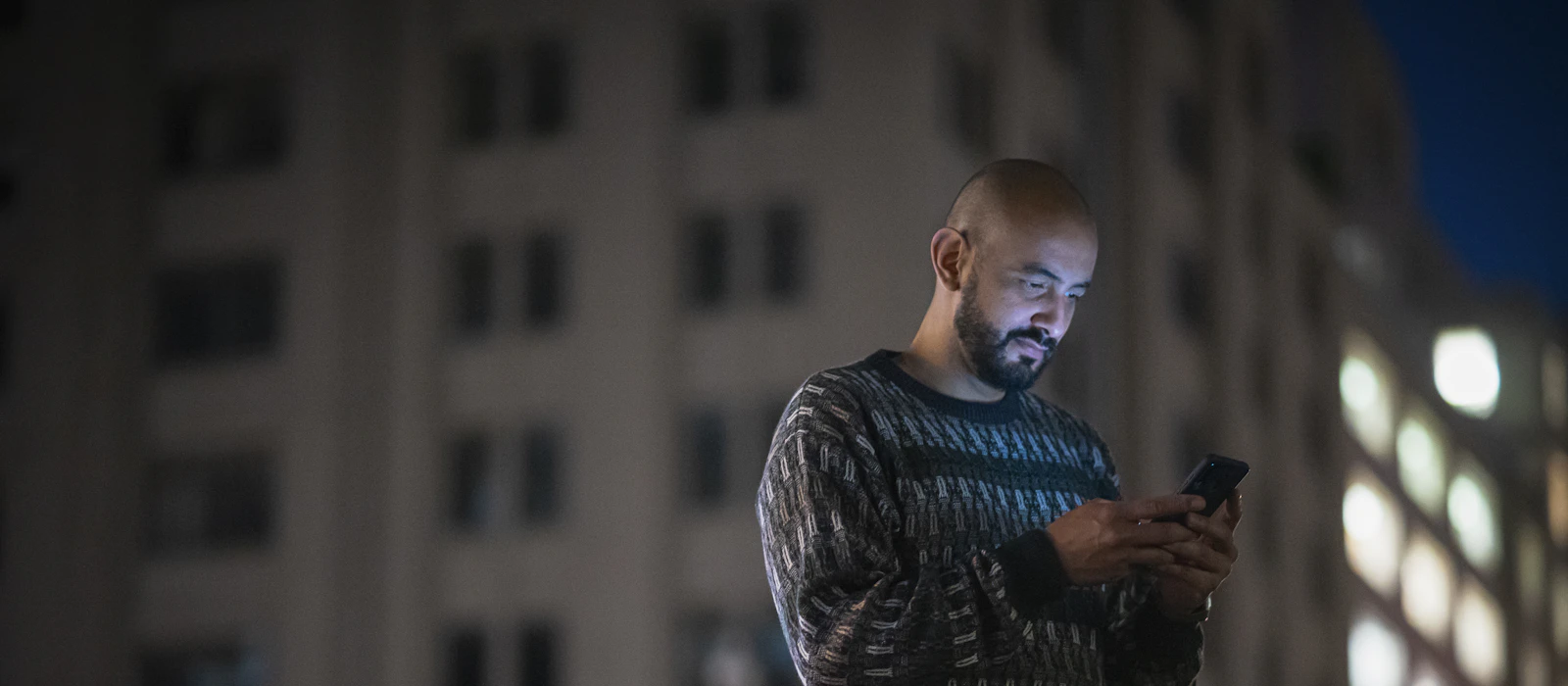 Man checking his smartphone outdoors at night with city buildings behind him